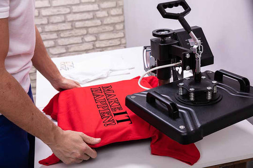 DTF Transfers vs Screen Printing article header featuring a red MAKE IT HAPPEN t-shirt being prepared on a heat press machine illustrating durability and cost comparison between DTF transfer and screen printing methods